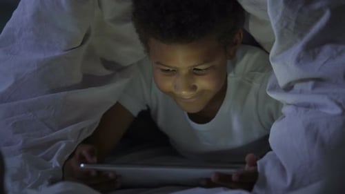 Petit enfant regardant la tablette secrètement la nuit, couvert de tête dans une couverture