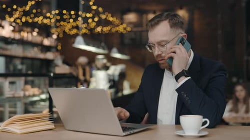 Businessman Working at Cafe Sitting with Laptop Holding Smartphone Talking