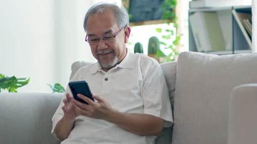 Senior Man Smiling Using Smartphone on Couch Indoors