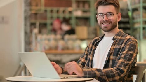 Young Man with Laptop Smiling at Camera in Cafe