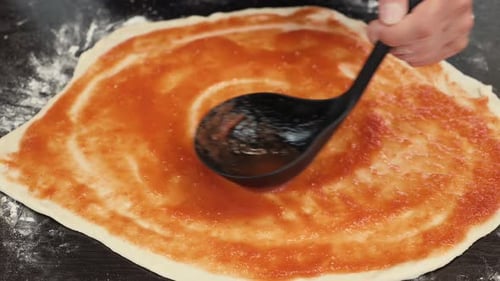 Female baker chef spreading tomato sauce on pizza dough, making pizza base