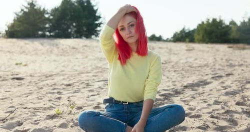 A Beautiful Serious Girl is Sitting on the Sand Outside on the Beach Pink Hair is Blown Away By the