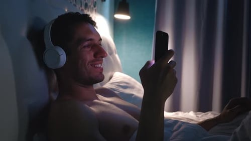 Smiling Young Man in Headphones Surfing the Internet and Listening Music with a Smartphone Before