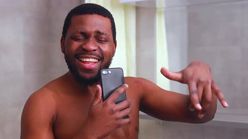 Attractive Young Cheerful Man Singing While Washing in the Shower Holding Phone
