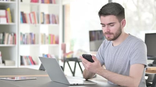 Junger Mann benutzt Smartphone und Laptop im Büro