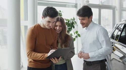 Young Man Signing Purchase Agreement at Automobile Showroom Shaking Hands with Sales Agent to