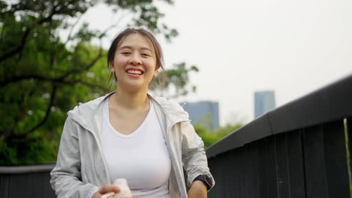 4K Asian woman drinking water from a bottle while jogging at public park in the morning.