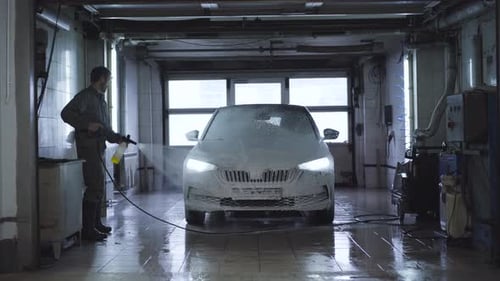 Wide Shot of Worker Washing Vehicle in Car Wash. Caucasian Employee Using High-pressure Washer at