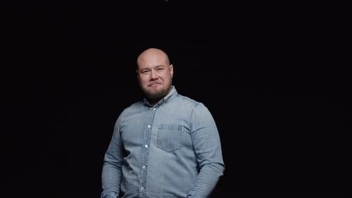 A bald man with a beard makes a surprised face and shakes his head affirmatively against a black