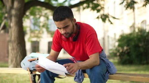Exhausted Young Male Student with Paperwork Holding Head in Hands Sitting on Bench Outdoors on Sunny