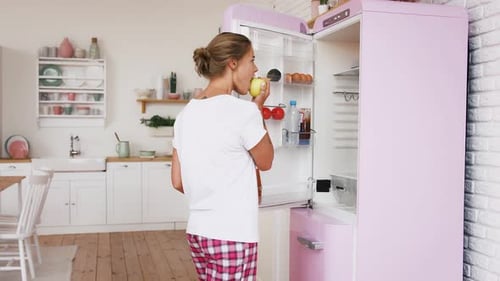 Young Woman Eats Apple, Grabs Lettuce From Refrigerator