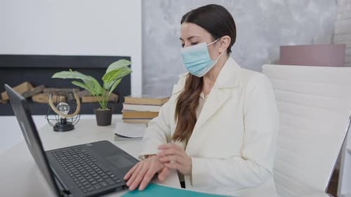 Side View of Exhausted Ill Young Woman in Covid19 Face Mask Sitting at Table in Office Coughing and