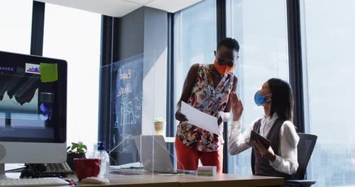 Diverse female office colleagues wearing face masks discussing at modern office