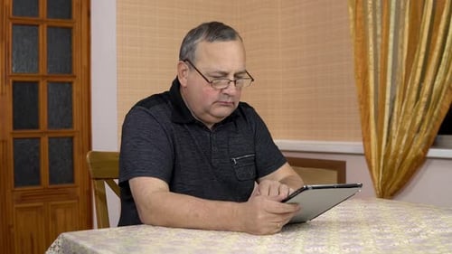 Serious Man Uses a Tablet, Old Man Works with a Tablet While Sitting at the Table