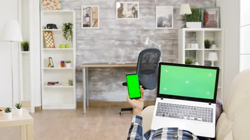 Man Lying on the Sofa Holding a Green Screen Laptop and Smartphone in His Hands