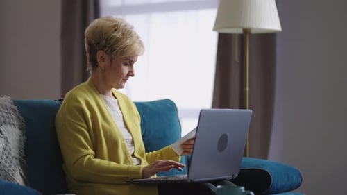 Woman Types on Laptop Holding Paper at Home