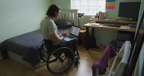 Fokussierter kaukasischer behinderter Mann im Rollstuhl mit Laptop im Schlafzimmer