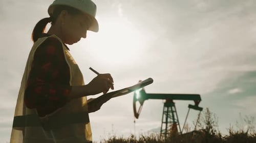 Silhouette Ingenieurin schreibt auf Klemmbrett im Ölfeld, Frau trägt weißen Helm und arbeitet