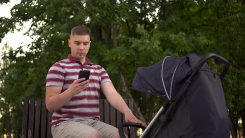 A Young Dad with a Phone in His Hand Cradles a Child in a Stroller