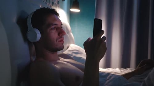 Young Man in Headphones Surfing the Internet and Listening Music with a Smartphone Before Bed