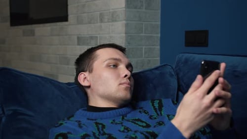 Close Up Young Caucasian Man Uses Smartphone While Laying on a Sofa at Home. Man Browses Through