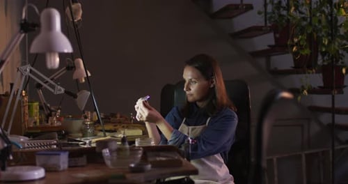 Profile of focused caucasian female jeweller sitting at desk, making jewelry in workshop