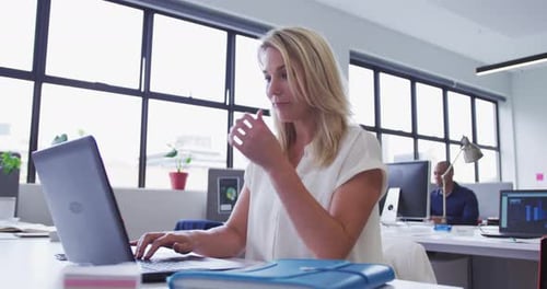 Caucasian businesswoman sitting using laptop in office with coworker in background