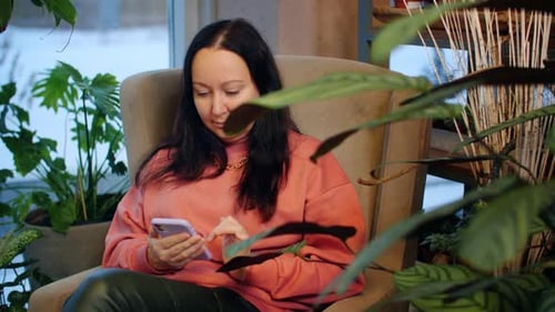 Relaxed Woman Using Mobile Phone in Soft Chair in Indoor Garden