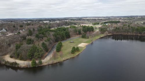 Luftaufnahme vom See zum Park mit Wald, einem Unterschlupf, offenem Feld und einem kleinen leeren Strand.