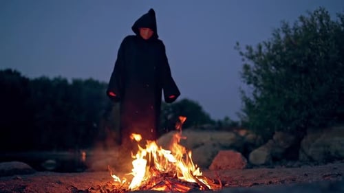Witch in black spooky costume. Scary wizard in cloak with hood doing shamanism over the burning fire