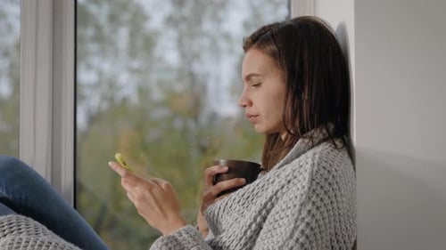 Woman Drinking Coffee and Using Phone by Window