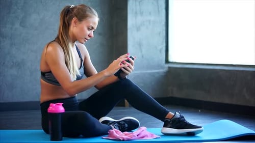 Woman in the Gym with Smartphone. She Sitting on the Mat and Using Smartphone