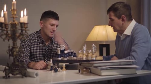 Cheerful Caucasian Son and Father Playing Chess in the Evening at Home. Adult Man Spending Free Time