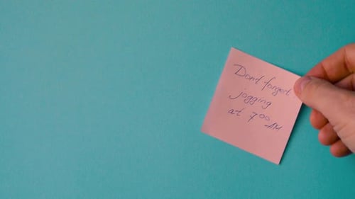 Male Hand is Pasting a Sticker with the Words Jogging at 7 AM Written in a Pen on a Blue Background