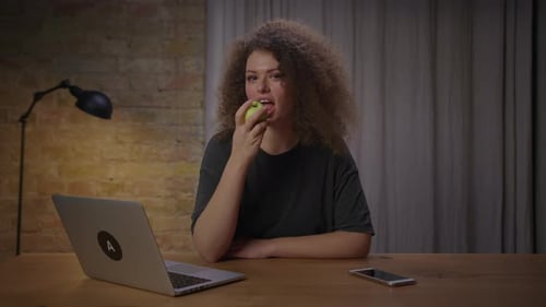 Young Woman Eating Green Apple Sitting at Table with Laptop at Home and Looking at Camera
