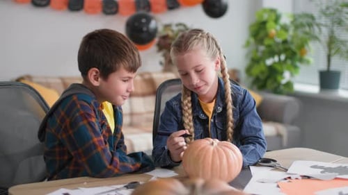 Children with Felttip Pen Draw a Face on a Pumpkin Against the Background of a Garland and Balloons