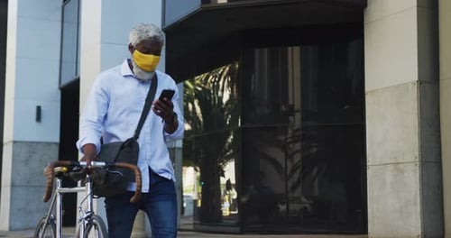 Hombre mayor afroamericano con máscara facial con bicicleta usando un teléfono inteligente en un parque Corporativa