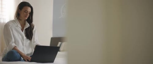 Eine junge Frau tippt auf ihrem Laptop, während sie im Bett sitzt und von zu Hause aus arbeitet