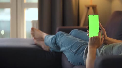 Woman at Home Lying on a Sofa and Using Smartphone with Green Mock-up Screen in Vertical Mode. Girl