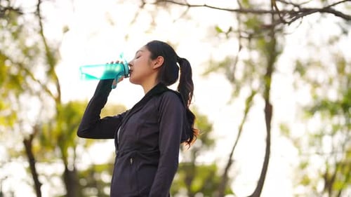 4K Asian woman drinking water from a bottle while jogging at public park in the morning.