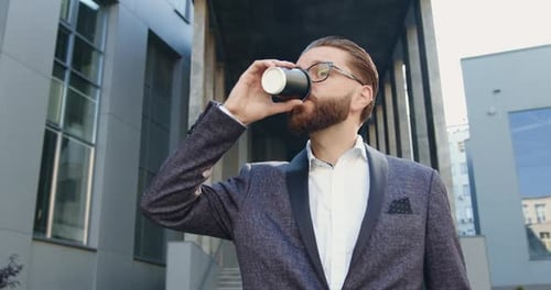 Man with Well-Groomed Beard in Glasses which Drinking Coffee Near Modern Urban Office Building