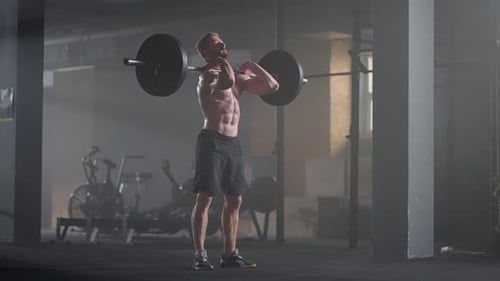 Shirtless Man Squats with Barbell in Misty Gym