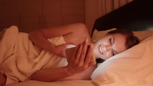 Woman Smiling Looking at Mobile Device in Bed