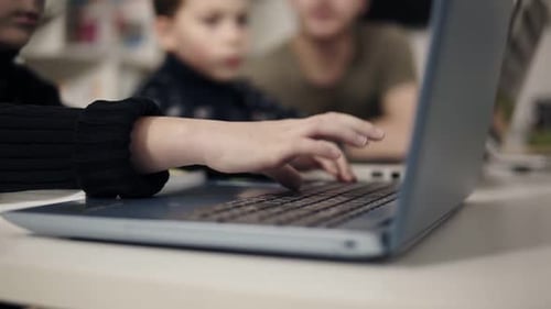 Kid's Arms Typing Somethingon Laptop Keyboard with Two Unrecognizable Figures Sitting on the