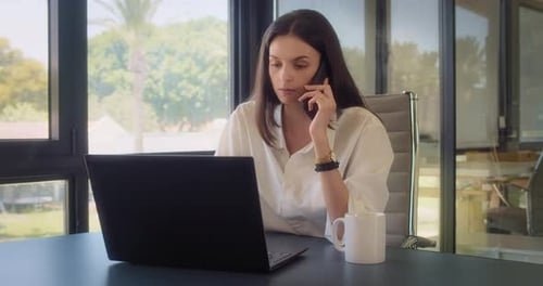 A businesswoman having a drink while talking on the phone at the office with big windows.