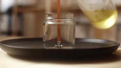 Closeup Front View of Unrecognizable Female Artisan Pouring Melted Wax Into Glass Vessel with Wick