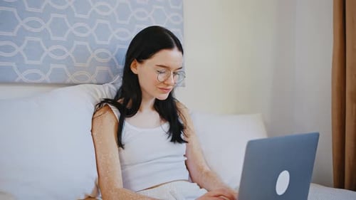 Verkleinerungsaufnahme einer Frau mit Vitiligo-Pigmentierung beim Tippen auf Laptop, die zu Hause langsam auf dem Bett sitzt