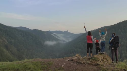 A Happy Family with Three Children Cheerfully Raise Their Hands Standing on the Top of the Mountain