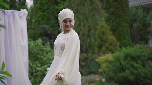 Joyful Smiling Middle Eastern Bride in White Wedding Dress and Hijab Spinning in Summer Garden at
