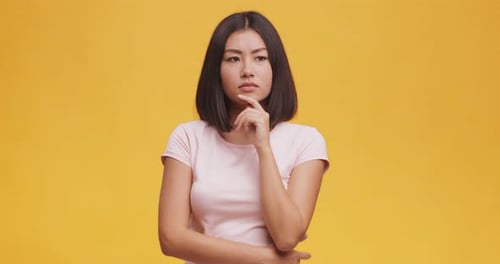Young Thoughtful Asian Woman Pondering About Something, Touching Her Chin, Orange Studio Background